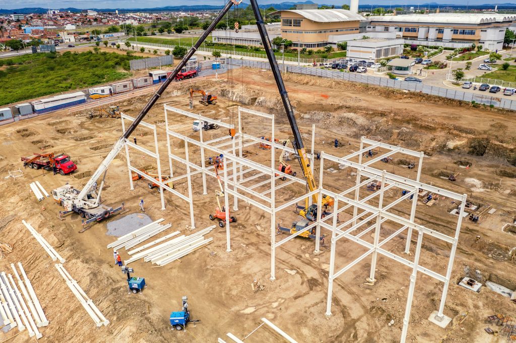 Vista aérea de canteiro de obras com guindastes e montagem de estrutura metálica.