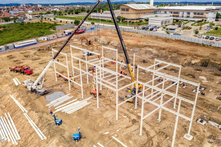Vista aérea de canteiro de obras com guindastes e montagem de estrutura metálica.